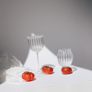 Dos copas de cristal transparente con relieve vertical junto a tomates sobre papel blanco, fondo con gradiente de luz y sombra