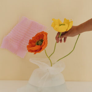Dos flores de amapola —una naranja y otra amarilla— en tallos curvados colocadas dentro de una bolsa plástica con agua, sobre una superficie blanca y fondo beige, mano sosteniendo rama amarilla y tapete rosa en el fondo