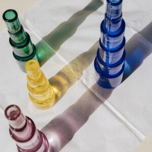 Varias botellas/jarras de vidrio translúcido en verde, amarillo, azul y rosa dispuestas sobre papel blanco, con sombras alargadas