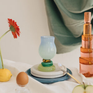 Mesa decorada con copa bicolor apoyada sobre platos, cucharilla dorada, flor gerbera roja en limón, botella de vidrio ámbar escalonada y manzana partida, fondo con tela verde